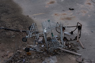 A broken and overturned shopping cart lies amidst scattered debris and dried leaves on a worn concrete surface. The surroundings appear neglected, with rusted metal parts and various trash pieces strewn about.