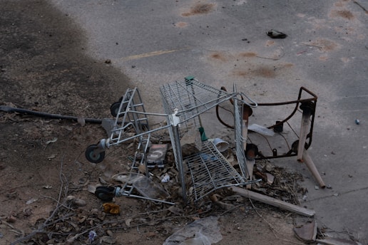 A broken and overturned shopping cart lies amidst scattered debris and dried leaves on a worn concrete surface. The surroundings appear neglected, with rusted metal parts and various trash pieces strewn about.