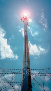 A tall telecommunications tower extends upwards towards the sky, with a bright light or sun at its peak. The sky is mostly clear with some fluffy clouds. The foreground features barbed wire, indicative of a fenced or restricted area.