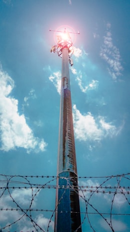 A tall telecommunications tower extends upwards towards the sky, with a bright light or sun at its peak. The sky is mostly clear with some fluffy clouds. The foreground features barbed wire, indicative of a fenced or restricted area.