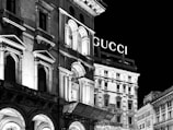 A nighttime view of an illuminated urban scene with classical architectural elements. The facade of a building features arched windows, decorative columns, and detailed cornices. A prominent sign displaying 'GUCCI' is visible atop one of the buildings in the background.