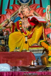 A statue of the Hindu deity Ganesha is adorned with vibrant yellow and red robes, sitting on a decorated throne. The figure holds a book and prayer beads, surrounded by offerings such as flowers and a lit oil lamp. Intricate patterns and floral garlands add to the ornate appearance.