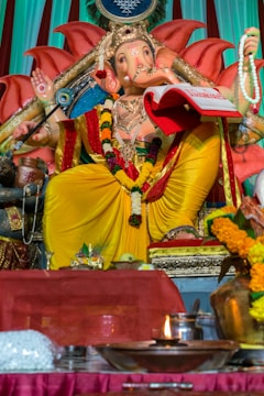 A statue of the Hindu deity Ganesha is adorned with vibrant yellow and red robes, sitting on a decorated throne. The figure holds a book and prayer beads, surrounded by offerings such as flowers and a lit oil lamp. Intricate patterns and floral garlands add to the ornate appearance.