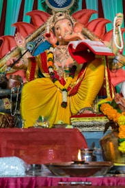 A statue of the Hindu deity Ganesha is adorned with vibrant yellow and red robes, sitting on a decorated throne. The figure holds a book and prayer beads, surrounded by offerings such as flowers and a lit oil lamp. Intricate patterns and floral garlands add to the ornate appearance.