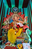 A large, ornate statue of Ganesha, the Hindu deity with an elephant head, is adorned with colorful garlands and traditional attire. The setting includes a vivid backdrop with vibrant colors and intricate patterns. Various offerings are placed at the base, including fruit and decorative items. The scene is rich with cultural symbolism and artistry.