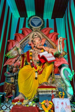 A large, ornate statue of Ganesha, the Hindu deity with an elephant head, is adorned with colorful garlands and traditional attire. The setting includes a vivid backdrop with vibrant colors and intricate patterns. Various offerings are placed at the base, including fruit and decorative items. The scene is rich with cultural symbolism and artistry.