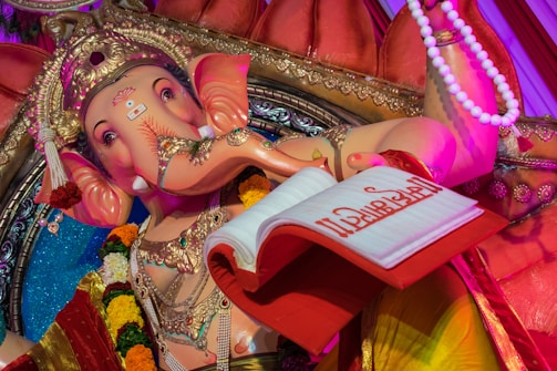 A detailed statue depicting a Hindu deity with an elephant head, adorned with an ornate headdress and intricate jewelry. The figure is holding a red book with gold writing and is surrounded by vibrant garlands of flowers. The setting features vivid colors and decorative elements.