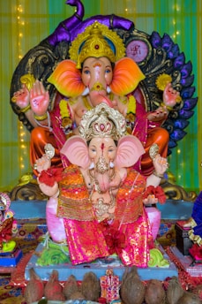 The image features two intricately decorated statues of Lord Ganesha, a widely worshipped Hindu deity with an elephant's head. The statues are adorned with vibrant garments and jewelry. The larger statue is seated in the background with a colorful halo around it, while the smaller statue is seated in front. The setting includes other small religious artifacts and coconuts placed at the base of the statues, indicating a possible religious or festive setup.