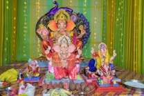 A vibrant and colorful display showcases multiple statues of Lord Ganesha, each adorned with ornate decorations and intricate garments. The background features green and yellow drapes with a string of lights, enhancing the festive ambiance. Various offerings, including coconuts and flowers, are arranged in front of the statues.