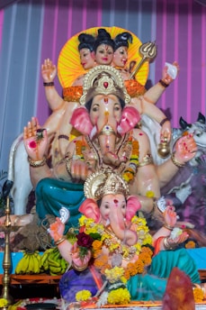 A detailed statue of Hindu deities, with multiple figures intricately adorned. The foremost figure is a representation of an elephant-headed deity, surrounded by colorful garlands and ornate jewelry. Behind, a multi-faced deity holding various symbolic items is depicted. The background features a striped pattern with a bright halo effect.