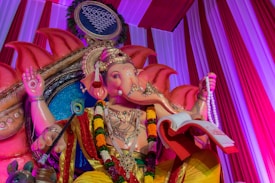 A vibrant and intricately decorated statue of the Hindu deity Ganesha, adorned with colorful garlands and traditional ornaments. The statue is set against a backdrop of red and purple drapery, featuring detailed carvings. Ganesha holds a book and a rosary in its hands.