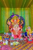 A brightly decorated display featuring a large statue of the Hindu god Ganesha, adorned in colorful clothing and ornaments. The background is lined with green and yellow drapes and illuminated with strings of lights. In front of the main statue are smaller statues of Ganesha and various offerings including coconuts, bananas, and other ritual items.