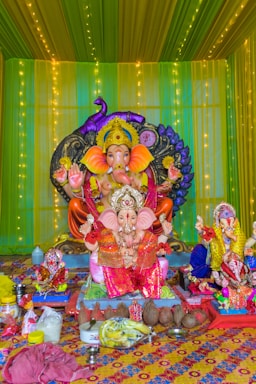 A brightly decorated display featuring a large statue of the Hindu god Ganesha, adorned in colorful clothing and ornaments. The background is lined with green and yellow drapes and illuminated with strings of lights. In front of the main statue are smaller statues of Ganesha and various offerings including coconuts, bananas, and other ritual items.