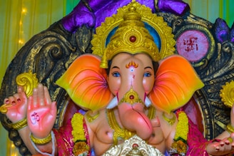 A colorful statue of Ganesha, the Hindu deity, with vibrant orange and pink hues on the ears and intricate gold embellishments on the head and jewelry. The hands are raised with religious symbols, and the statue is adorned with garlands. The backdrop features decorative elements and patterns.