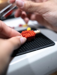 Hands are assembling small red and yellow pieces on a black base, likely engaging in a crafting or building activity.