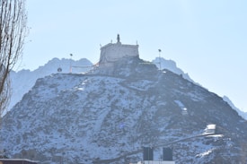 A white structure resembling a monastery or temple is situated atop a snow-dusted hill or mountain. The peaks of the mountains in the background are visible under a clear blue sky. There are prayer flags strung near the structure, and some foliage is present on the left side.