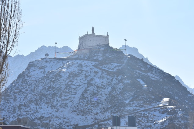 A white structure resembling a monastery or temple is situated atop a snow-dusted hill or mountain. The peaks of the mountains in the background are visible under a clear blue sky. There are prayer flags strung near the structure, and some foliage is present on the left side.