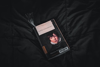 A copy of David Copperfield by Charles Dickens lies on a dark, textured fabric surface. The book cover features an illustrated portrait of a young person, with a black, white, and red color scheme. A tassel bookmark protrudes from the top of the book, adding texture and dimension.