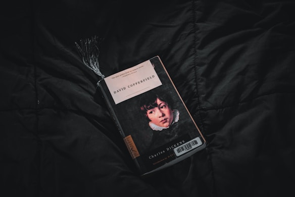 A copy of David Copperfield by Charles Dickens lies on a dark, textured fabric surface. The book cover features an illustrated portrait of a young person, with a black, white, and red color scheme. A tassel bookmark protrudes from the top of the book, adding texture and dimension.
