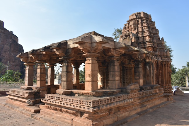 An ancient stone temple featuring intricate carvings and multiple stone pillars. The architecture includes detailed friezes and a tiered tower that rises above the structure. Surrounding the temple are natural elements such as trees and a rocky landscape.