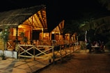 A series of thatched-roof wooden cottages are warmly lit and stand in a row. Two people sit at a table outside near the cottages, and the area is surrounded by trees.