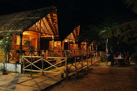 A series of thatched-roof wooden cottages are warmly lit and stand in a row. Two people sit at a table outside near the cottages, and the area is surrounded by trees.