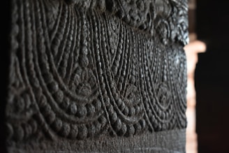 Intricate stone carving featuring detailed, symmetrical patterns with a rich texture. The grooves form curving, wave-like designs that suggest traditional or historical artistry. Dim lighting highlights the relief with shadows, accentuating its depth.