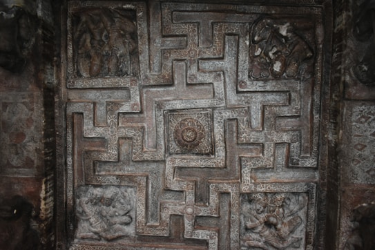 An intricately carved stone relief displaying a symmetrical maze pattern with detailed, artistic engravings. The central focus is a circular medallion surrounded by geometric shapes, possibly representing a labyrinth. Surrounding panels depict figures in dynamic poses, suggesting mythological or cultural significance.