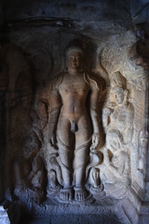 An ancient stone carving depicting a standing figure surrounded by smaller figures on a textured rock surface. The central figure appears to be adorned with simple garments and exhibits a calm expression. The surrounding figures are intricately carved and seem to be in various poses, adding to the artistic complexity of the scene.