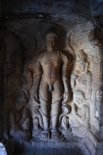 An ancient stone carving depicting a standing figure surrounded by smaller figures on a textured rock surface. The central figure appears to be adorned with simple garments and exhibits a calm expression. The surrounding figures are intricately carved and seem to be in various poses, adding to the artistic complexity of the scene.