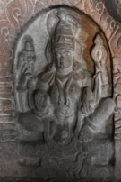 A stone sculpture depicting a seated deity, possibly from ancient or classical art. The figure has multiple arms, holding various symbolic objects. The carving is detailed, showing intricate patterns and designs surrounding the figure.