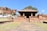 A historic stone temple with classical Indian architecture, set against a backdrop of rocky cliffs and a clear blue sky. The temple features ornamental pillars and a rectangular roof, surrounded by a grassy area with a paved path leading up to it. Nearby structures and a tree are visible, enhancing the ancient and serene setting.
