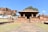 A historic stone temple with classical Indian architecture, set against a backdrop of rocky cliffs and a clear blue sky. The temple features ornamental pillars and a rectangular roof, surrounded by a grassy area with a paved path leading up to it. Nearby structures and a tree are visible, enhancing the ancient and serene setting.