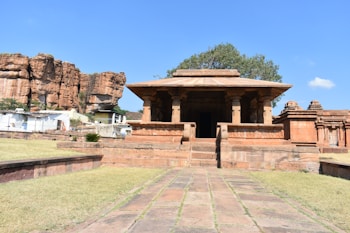 A historic stone temple with classical Indian architecture, set against a backdrop of rocky cliffs and a clear blue sky. The temple features ornamental pillars and a rectangular roof, surrounded by a grassy area with a paved path leading up to it. Nearby structures and a tree are visible, enhancing the ancient and serene setting.
