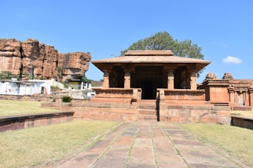 A historic stone temple with classical Indian architecture, set against a backdrop of rocky cliffs and a clear blue sky. The temple features ornamental pillars and a rectangular roof, surrounded by a grassy area with a paved path leading up to it. Nearby structures and a tree are visible, enhancing the ancient and serene setting.