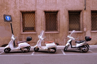 Three scooters are parked in a row next to a weathered, brownish wall with barred windows. Each scooter has a windshield and is positioned in a marked parking space. A nearby blue road sign indicates one-way traffic, and the setting appears to be urban.