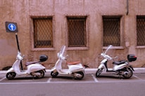 Three scooters are parked in a row next to a weathered, brownish wall with barred windows. Each scooter has a windshield and is positioned in a marked parking space. A nearby blue road sign indicates one-way traffic, and the setting appears to be urban.