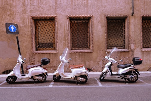 Three scooters are parked in a row next to a weathered, brownish wall with barred windows. Each scooter has a windshield and is positioned in a marked parking space. A nearby blue road sign indicates one-way traffic, and the setting appears to be urban.