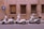 Three scooters are parked in a row next to a weathered, brownish wall with barred windows. Each scooter has a windshield and is positioned in a marked parking space. A nearby blue road sign indicates one-way traffic, and the setting appears to be urban.