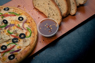 A freshly baked pizza with a variety of toppings including black olives, green and red peppers, mushrooms, and corn. Next to the pizza, there is a small container of dipping sauce with chili flakes. In the background, thick slices of herb bread are placed on a wooden board.