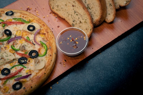 A freshly baked pizza with a variety of toppings including black olives, green and red peppers, mushrooms, and corn. Next to the pizza, there is a small container of dipping sauce with chili flakes. In the background, thick slices of herb bread are placed on a wooden board.