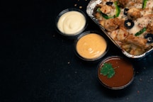 A foil container filled with a savory dish featuring diced meat, sliced black olives, and green bell peppers, accompanied by three small cups of sauces. The sauces range in color from creamy white, to a pale orange, and a rich reddish-brown garnished with a parsley leaf. The background is a dark surface.