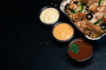 A foil container filled with a savory dish featuring diced meat, sliced black olives, and green bell peppers, accompanied by three small cups of sauces. The sauces range in color from creamy white, to a pale orange, and a rich reddish-brown garnished with a parsley leaf. The background is a dark surface.