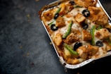 A close-up view of a foil container filled with baked food, featuring chunks of grilled chicken, sliced green peppers, black olives, and melted cheese. The dish has a crispy appearance with sprinkles of seasoning visible on top.