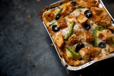 A close-up view of a foil container filled with baked food, featuring chunks of grilled chicken, sliced green peppers, black olives, and melted cheese. The dish has a crispy appearance with sprinkles of seasoning visible on top.