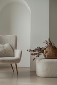 A cozy and elegant interior setup featuring a textured chair with slender legs, paired with a fluffy cushion. Next to it, a woven basket filled with dried decorative branches rests on a soft, matching ottoman. The scene is bathed in soft, neutral tones with a subtle archway in the background adding to the minimalist aesthetic.