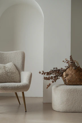 A cozy and elegant interior setup featuring a textured chair with slender legs, paired with a fluffy cushion. Next to it, a woven basket filled with dried decorative branches rests on a soft, matching ottoman. The scene is bathed in soft, neutral tones with a subtle archway in the background adding to the minimalist aesthetic.