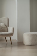A modern, minimalist room featuring a cozy armchair with a fluffy cushion and a matching round ottoman. Both furniture pieces have a soft, textured fabric in a light cream color. They are set against a light-colored wall with a unique architectural design.
