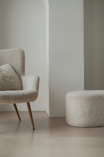 A modern, minimalist room featuring a cozy armchair with a fluffy cushion and a matching round ottoman. Both furniture pieces have a soft, textured fabric in a light cream color. They are set against a light-colored wall with a unique architectural design.
