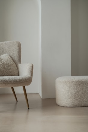 A modern, minimalist room featuring a cozy armchair with a fluffy cushion and a matching round ottoman. Both furniture pieces have a soft, textured fabric in a light cream color. They are set against a light-colored wall with a unique architectural design.